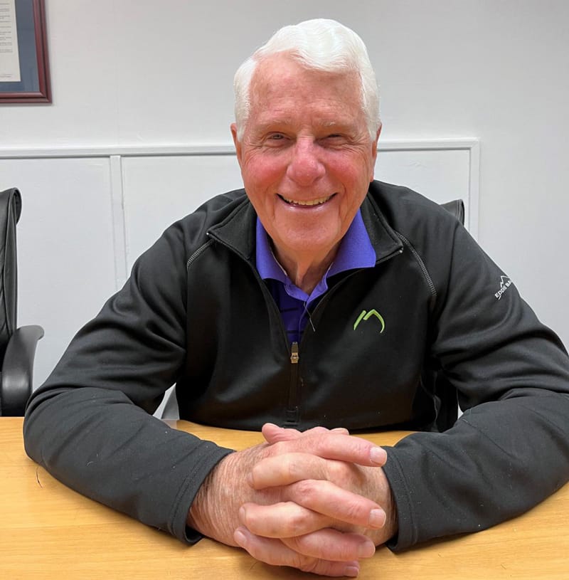 Jim Seeley smiles while sitting down with his hands clasped on the table