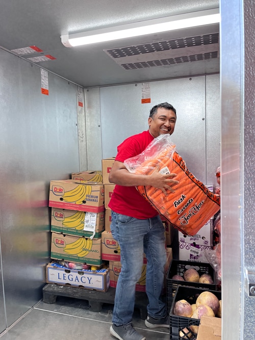Jose stands in the new walk-in fridge lifting a big bag of carrots and smiling