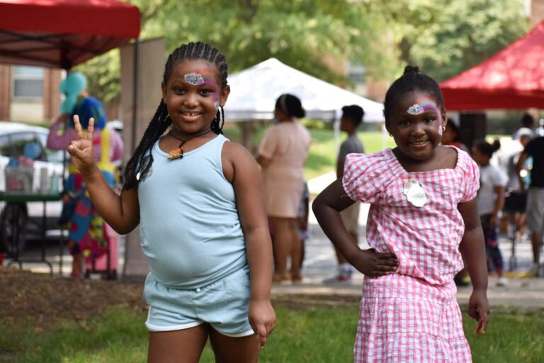 Two young girls at an event, red tents in the background, smiling. They have face paint and look like they're having a fun time.